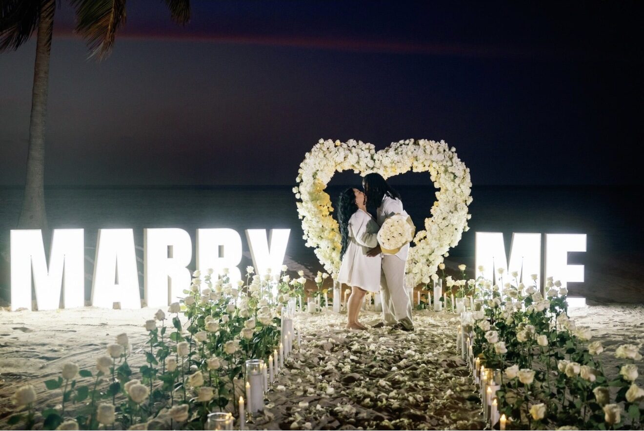 Couple kissing beneath a white flower heart arch beside illuminated MARRY ME letters on a Punta Cana beach at night