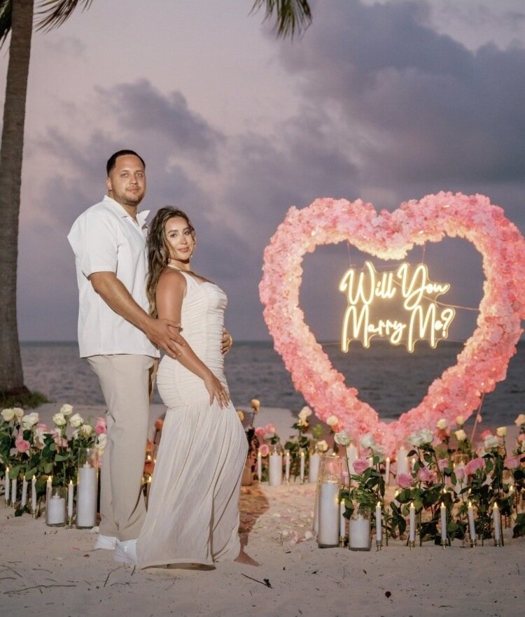 Couple embracing beside a pink rose heart arch reading 'Will You Marry Me' on a Punta Cana beach at dusk