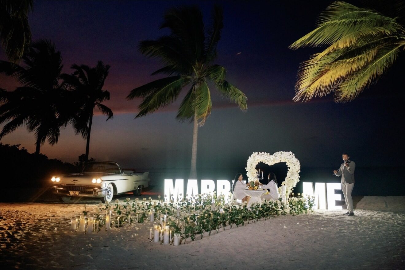 Vintage convertible and illuminated MARRY ME letters beside a white heart arch proposal setup at night in Punta Cana